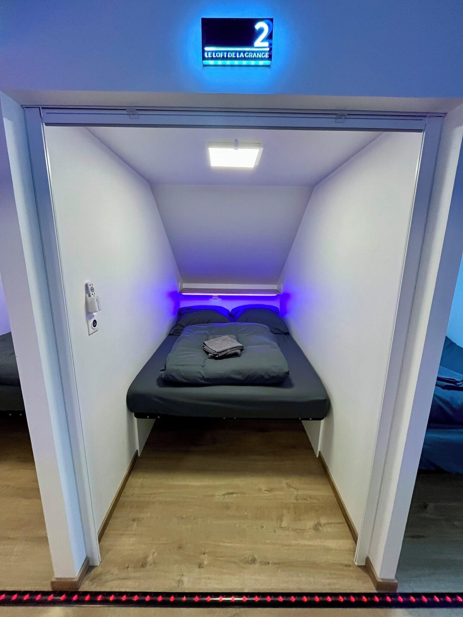 Une petite pièce fermée, avec un lit, une enseigne au-dessus indiquant « 2 Le Loft de la Grange », et un luminaire au plafond.