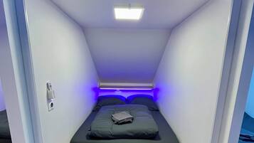 Une petite pièce fermée, avec un lit, une enseigne au-dessus indiquant « 2 Le Loft de la Grange », et un luminaire au plafond.