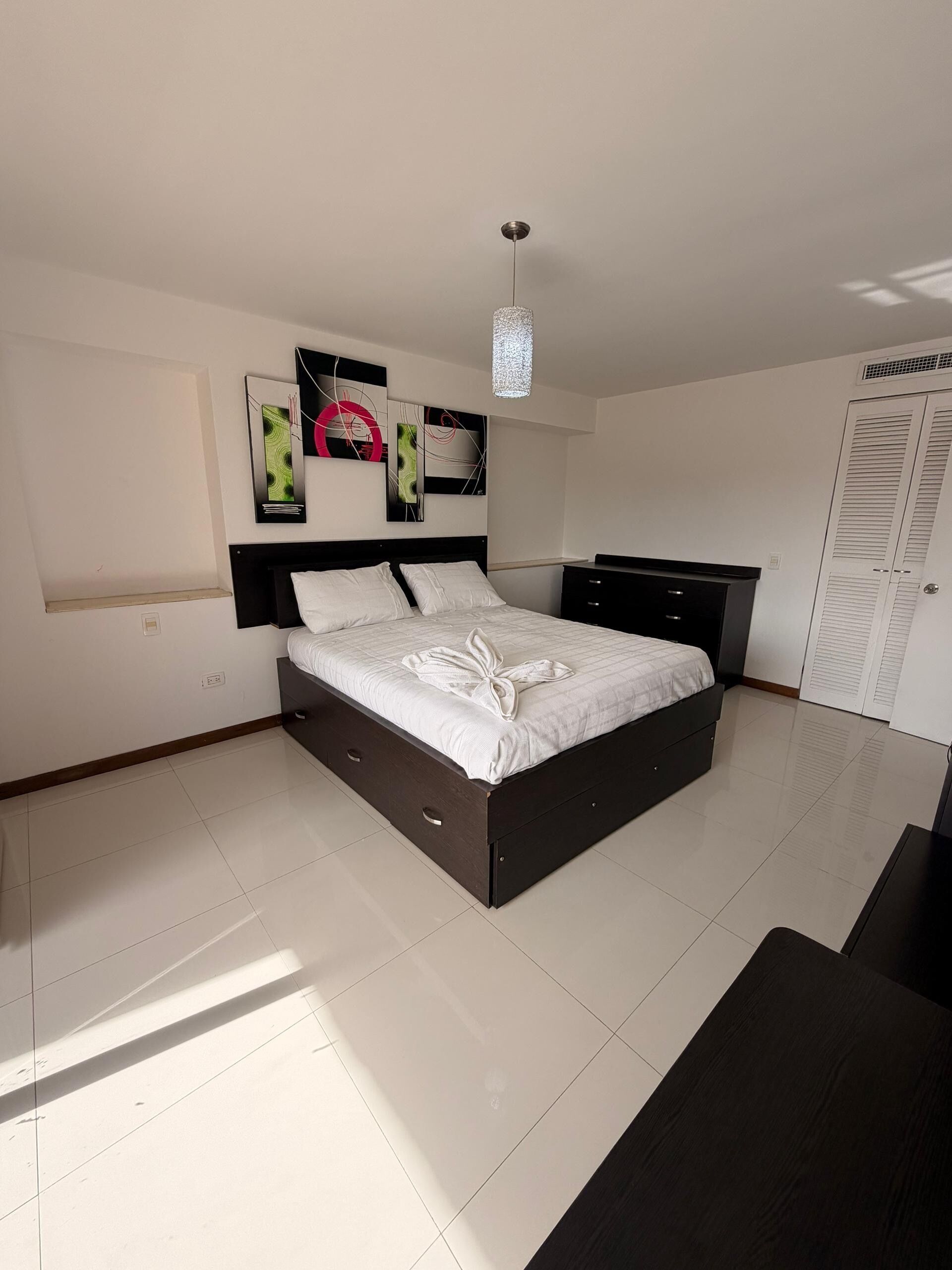 A modern bedroom with a large bed, a bedside table, a wardrobe, and abstract wall art.