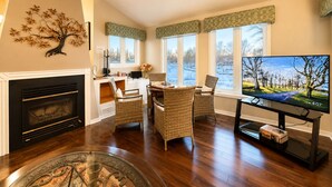 Una sala de estar con chimenea, muebles de mimbre, televisor y ventanas amplias que dan a un paisaje nevado.