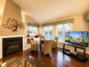 A living room with a fireplace, wicker furniture, a television, and large windows overlooking a snowy landscape. - Canalside summer cottage w/ river views, fire table & outdoor cushioned seating (Brechin)