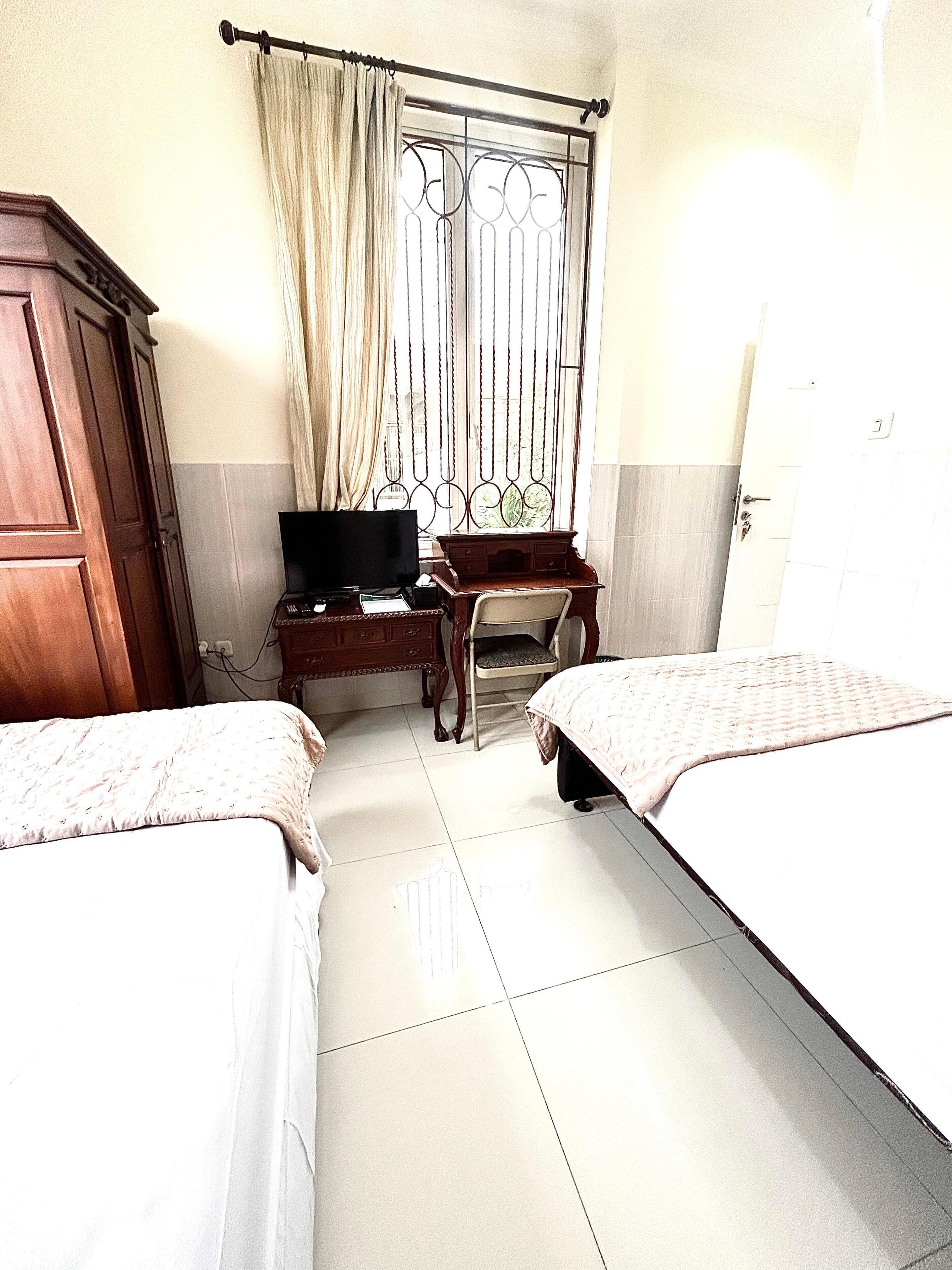 Habitación de hotel con dos camas, un escritorio con televisor, un armario y una ventana con barrotes.