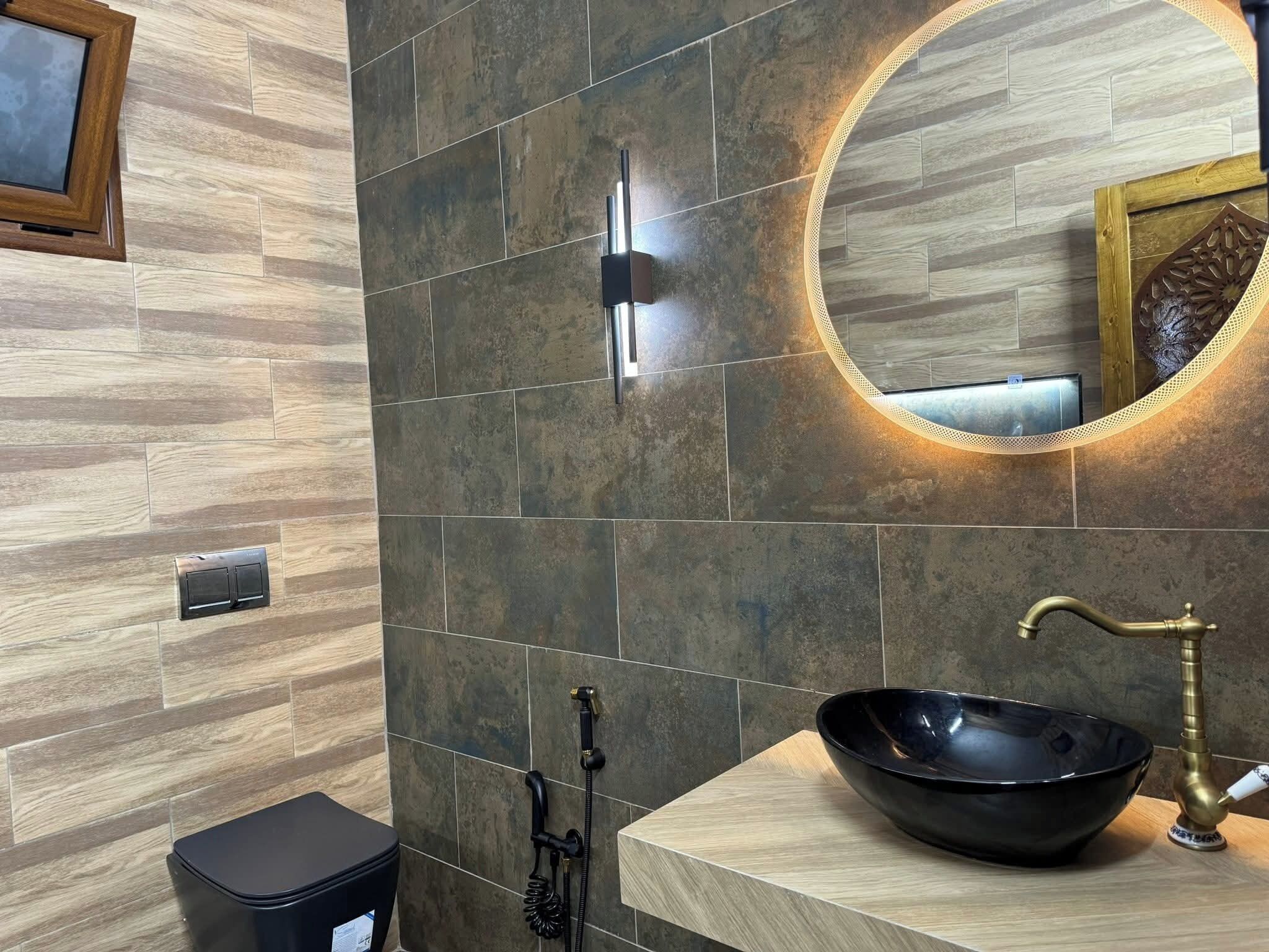 A bathroom with a wooden vanity, a black vessel sink, a brass faucet, a handheld showerhead, and a wall-mounted light fixture.