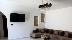 A spacious living room with a large sectional sofa, a coffee table, a flat-screen TV mounted on the wall, and a chandelier. - Very charming villa with swimming pool not overlooked, ideal for family or couple (Djerba Midun)