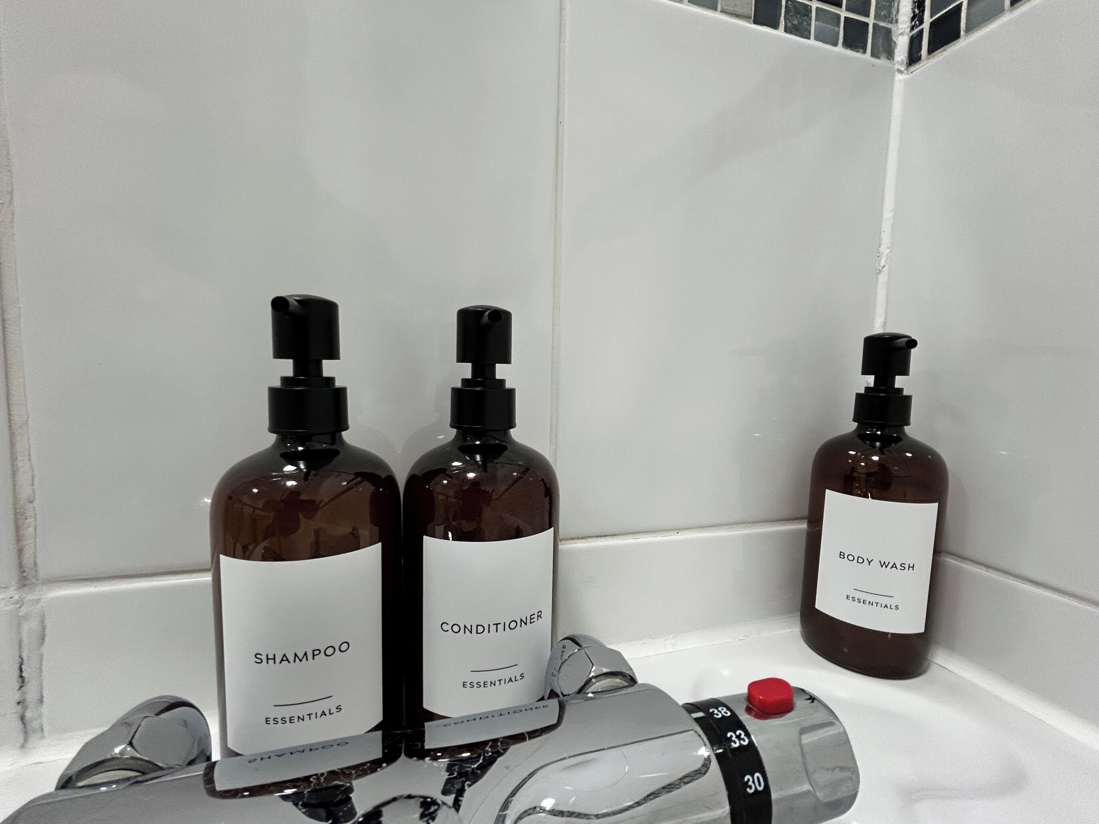 Un baño con pared de azulejos blancos, zona de ducha y tres botellas de artículos de aseo personal con las etiquetas 'Shampoo Essentials', 'Conditioner Essentials' y 'Body Wash Essentials'.