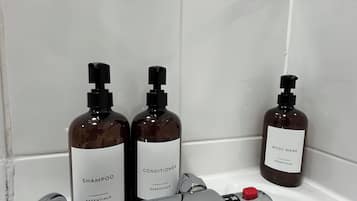 Un baño con pared de azulejos blancos, zona de ducha y tres botellas de artículos de aseo personal con las etiquetas 'Shampoo Essentials', 'Conditioner Essentials' y 'Body Wash Essentials'.