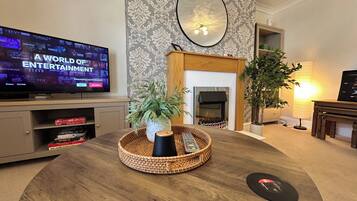 Una sala de estar con una televisión mostrando un servicio de transmisión, una chimenea y una mesa redonda de madera con una planta y una cesta.