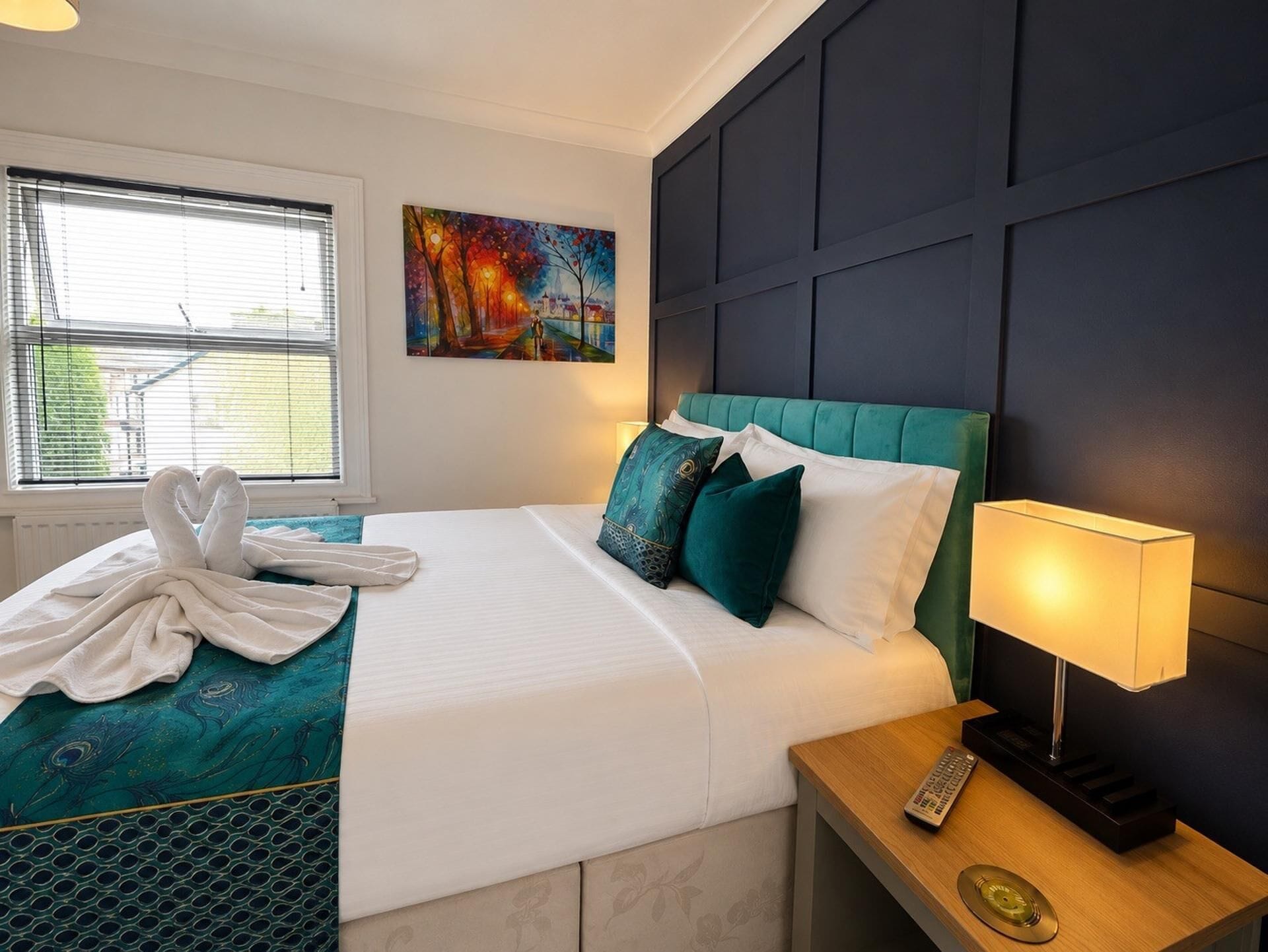 Habitación de hotel con cama, un cuadro, una ventana y una mesita de noche con lámpara y control remoto.