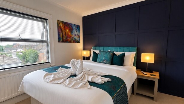 A hotel room with a large bed, a window with blinds, a painting on the wall, and two bedside lamps. - Character Bedford Apartment With Two Bedrooms (Bedford)