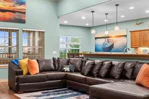 A living room with a sectional sofa, a kitchen area with pendant lights, and a painting of a boat on the wall. - Beautiful remodel, Game Room, Hot Tub, Pools, GOLF (Branson West)
