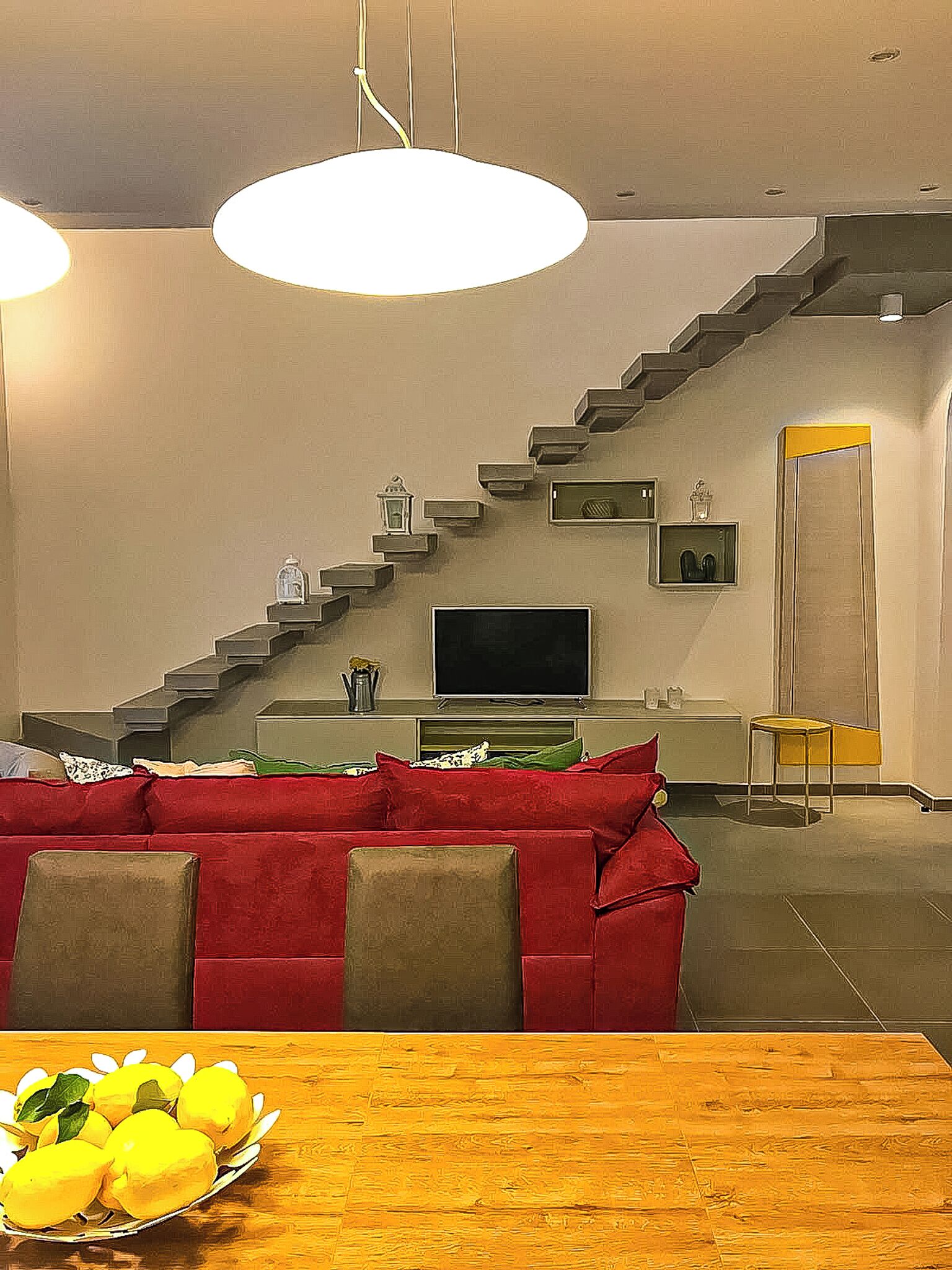 A modern living room with a red sofa, a wooden dining table with lemons, a staircase, and a TV.