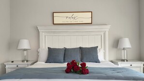 Un dormitorio con cabecera blanca, dos mesitas de noche con lámparas, cama con ropa de cama gris y un cartel enmarcado sobre la cama.