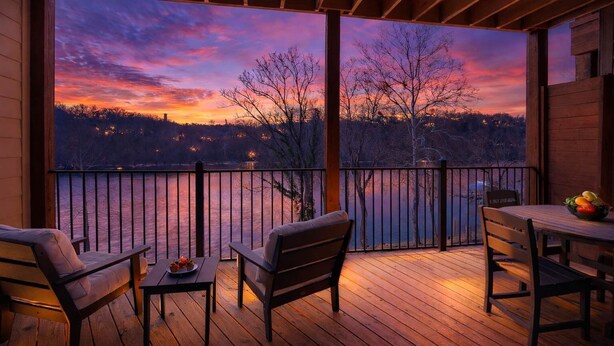 Una terraza de madera con vista a un lago al atardecer, una mesa con sillas y un cuenco de fruta.