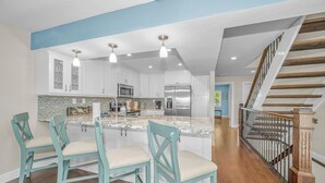 Une cuisine avec des meubles blancs, un réfrigérateur en acier inoxydable et un comptoir en granit. On y trouve trois chaises turquoise avec des coussins beige.