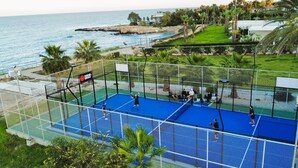 Ein Tennisplatz mit Netz, umzäunt und mit Blick auf eine Küstenlandschaft mit Schwimmbad und Grünflächen.