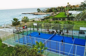 Ein Tennisplatz mit Netz, umzäunt und mit Blick auf eine Küstenlandschaft mit Schwimmbad und Grünflächen.