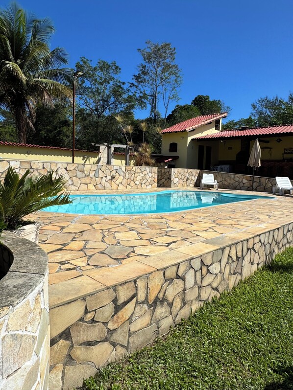 A swimming pool with stone tiles, a stone wall, and a building with a red roof in the background. - Fazendinha dos pássaros, your perfect refuge in Saquarema. (Saquarema)