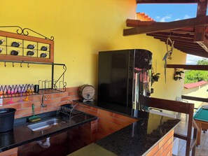 A kitchen with a sink, a hanging rack with utensils, a wooden barrel, and a refrigerator. - Fazendinha dos pássaros, your perfect refuge in Saquarema. (Saquarema)