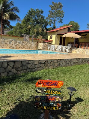 A pool area with a stone wall, a yellow building with a red roof, and a signpost with colorful directions. - Fazendinha dos pássaros, your perfect refuge in Saquarema. (Saquarema)