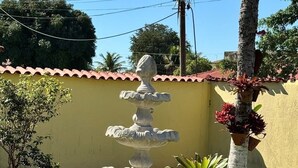 Uma fonte de pedra com vários níveis, rodeada por pedrinhas brancas, em um jardim gramado com um muro amarelo e palmeiras ao fundo.