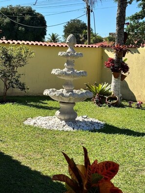 A stone fountain with multiple tiers, surrounded by white pebbles, in a grassy garden area with a yellow wall and palm trees in the background. - Fazendinha dos pássaros, your perfect refuge in Saquarema. (Saquarema)