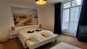 Ein Schlafzimmer mit Bett, Nachttisch, Lampe, einem Fenster mit Vorhängen und einem Bild an der Wand.