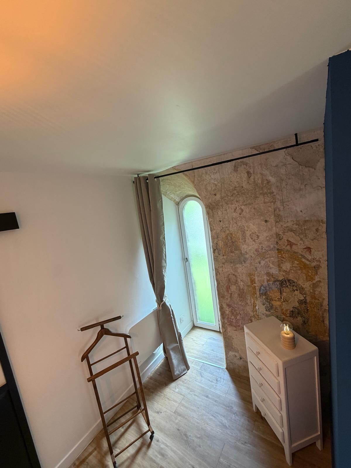 Ein Zimmer mit schrägen Wänden, einem hölzernen Kleiderbügel, einem weißen Schminktisch und einem kleinen Tisch mit Kerze.