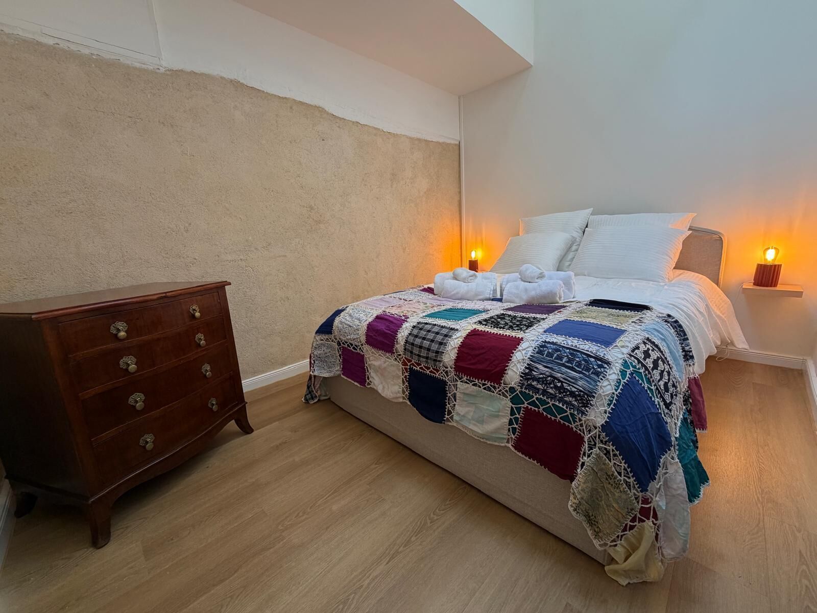 A bedroom with a wooden dresser, a quilted bedspread, two bedside lamps, and a wall-mounted shelf.