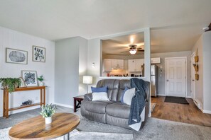 Ein Wohnzimmer mit grauem Sofa, einem Holztisch und einem kleinen Beistelltisch mit Pflanze. Durch einen offenen Durchgang ist eine Küche sichtbar.
