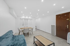 Ein Zimmer mit einem Bett, einem kleinen Tisch, einem Esstisch mit Stühlen, einer Holztür und einer weißen Wand.