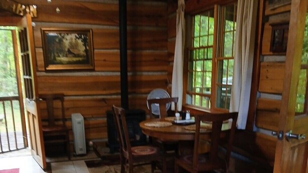A wooden cabin interior with a dining table, chairs, a fireplace, and a view of the outdoors through large windows. - Cozy 1-bedroom cabin in gorgeous Maggie Valley with AC (Maggie Valley)