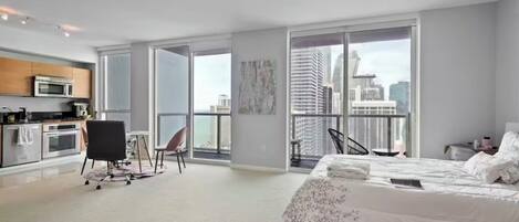 Une chambre moderne avec un grand lit, un bureau, une chaise et une vue sur le paysage urbain grâce à de grandes fenêtres.