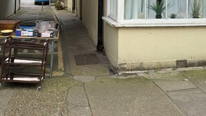 A narrow alleyway with a building on the right, a blue van in the distance, and a table with various items outside.