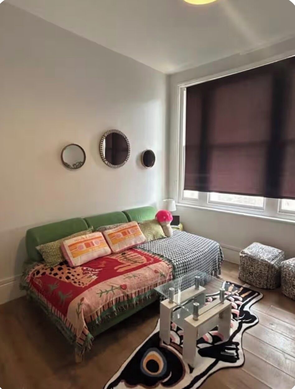 Una habitación con un sofá verde, una manta estampada, una mesa de centro de vidrio y un ventanal con persiana.