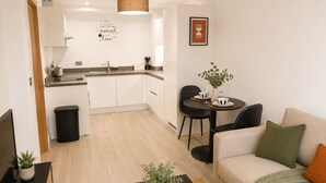 Eine moderne Küche mit weißen Schränken, ein Essbereich mit einem schwarzen Tisch und Stühlen, ein beiges Sofa mit einem grünen Kissen und eine kleine Pflanze auf einem schwarzen Tisch.