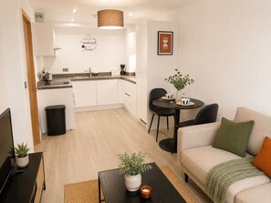 Eine moderne Küche mit weißen Schränken, ein Essbereich mit einem schwarzen Tisch und Stühlen, ein beiges Sofa mit einem grünen Kissen und eine kleine Pflanze auf einem schwarzen Tisch.