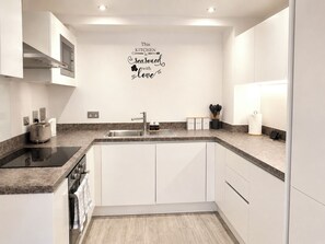 Eine moderne Küche mit weißen Schränken, einer Granit Arbeitsplatte, einem Edelstahlspülbecken und einer Mikrowelle. Eine Wanddeko zeigt den Schriftzug: 'Diese Küche ist mit Liebe zubereitet'.