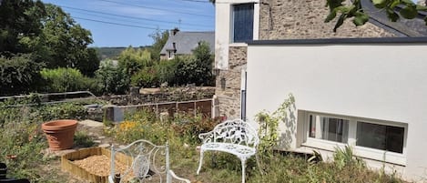 Un jardin avec un banc en métal blanc, un grand pot en terre cuite et un mur de pierre adjacent à un bâtiment doté d’une fenêtre.