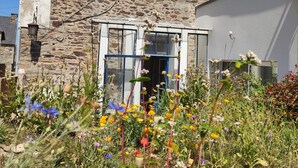 A stone house with a large window and a door, surrounded by a garden with various flowers.