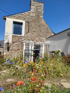 Ein Steinhaus mit großem Fenster und einer Tür, umgeben von einem Garten mit verschiedenen Blumen.