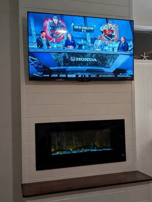Een televisie aan een wand bevestigd, waarop een ijshockeywedstrijd wordt getoond, met daaronder een open haard en een zeester decoratie.