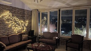A cozy living room with a brick wall, a chandelier, and a view of a cityscape at night. - Secluded Hilltop Villa Near Ancient Jarash — Privacy, Views & Pure Tranquility (Jerash)