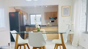 A modern kitchen with a glass dining table and chairs, a refrigerator, and a window with curtains. - 3 BEDROOMS AND TWO BATHROOMS, CLOSE TO DOWNTOWN, WITH 2 SWIMMING POOLS IN THE COMMON AREA. (Punta Cana)