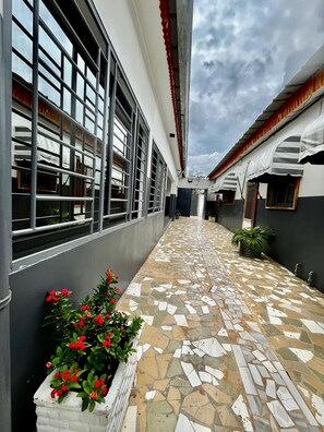 A corridor with tiled flooring, a white wall with a window, and a potted plant with red flowers. - 2-room apartment—living room and kitchen—5 minutes from the Institut National Polytechnique (Yamoussoukro)