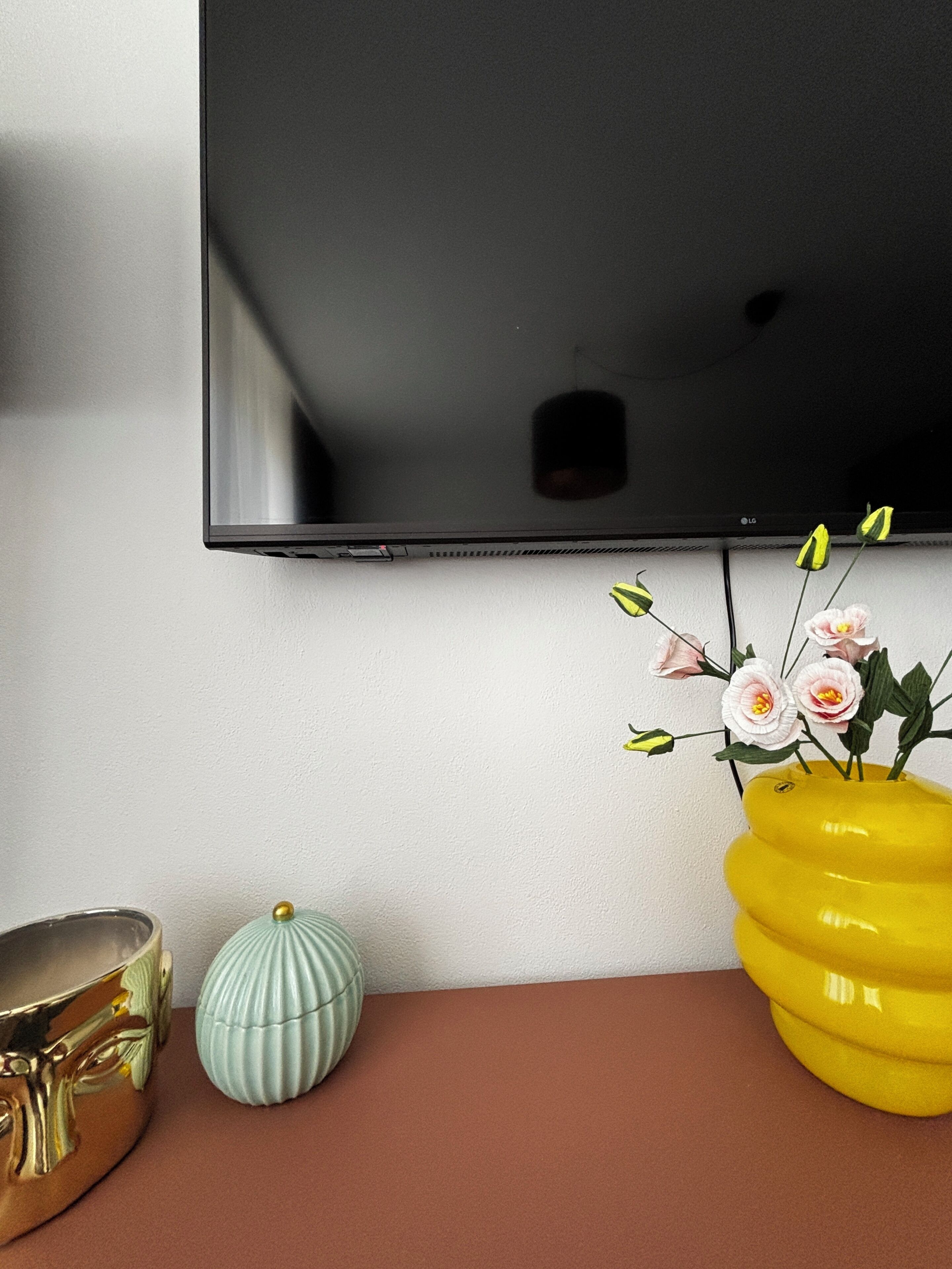 Ein Flachbildfernseher an der Wand, ein gelber Blumenvasen und zwei dekorative Vasen auf einem Tisch.