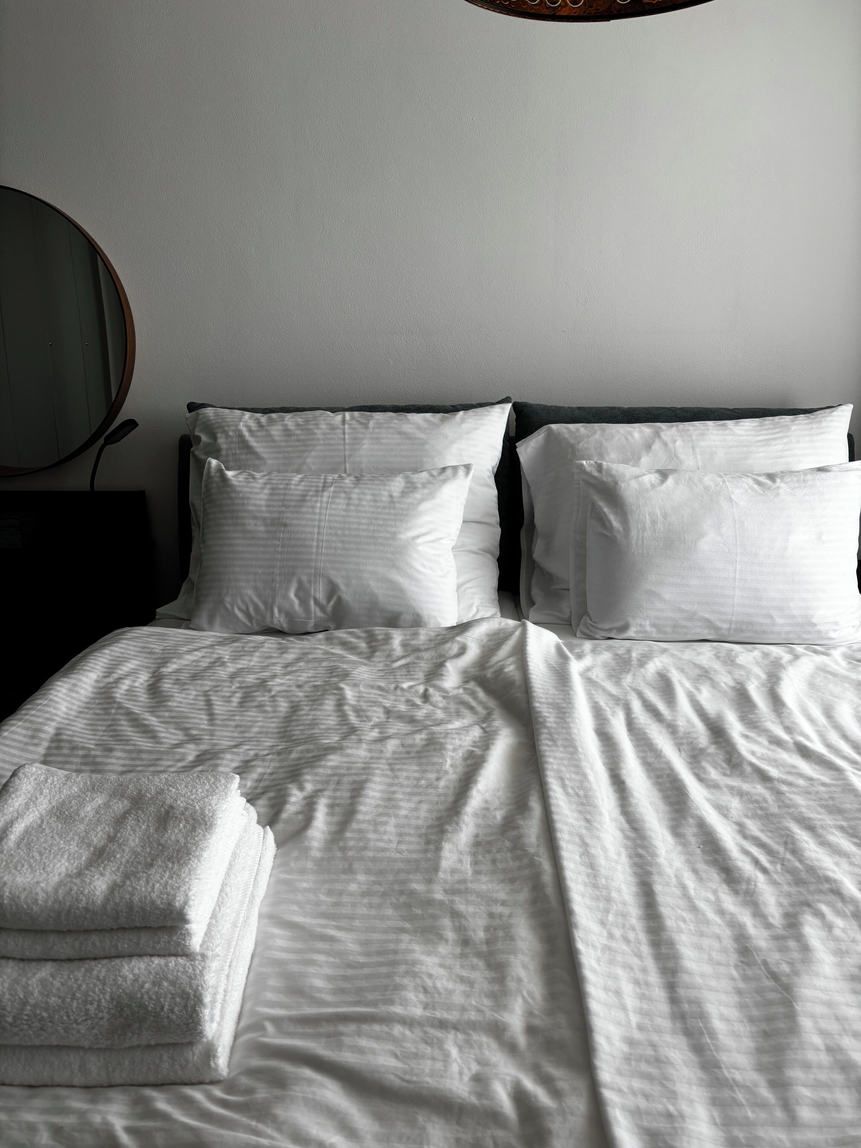 A neatly made bed with white linens and pillows, a round mirror on the wall, and a dark-colored nightstand.