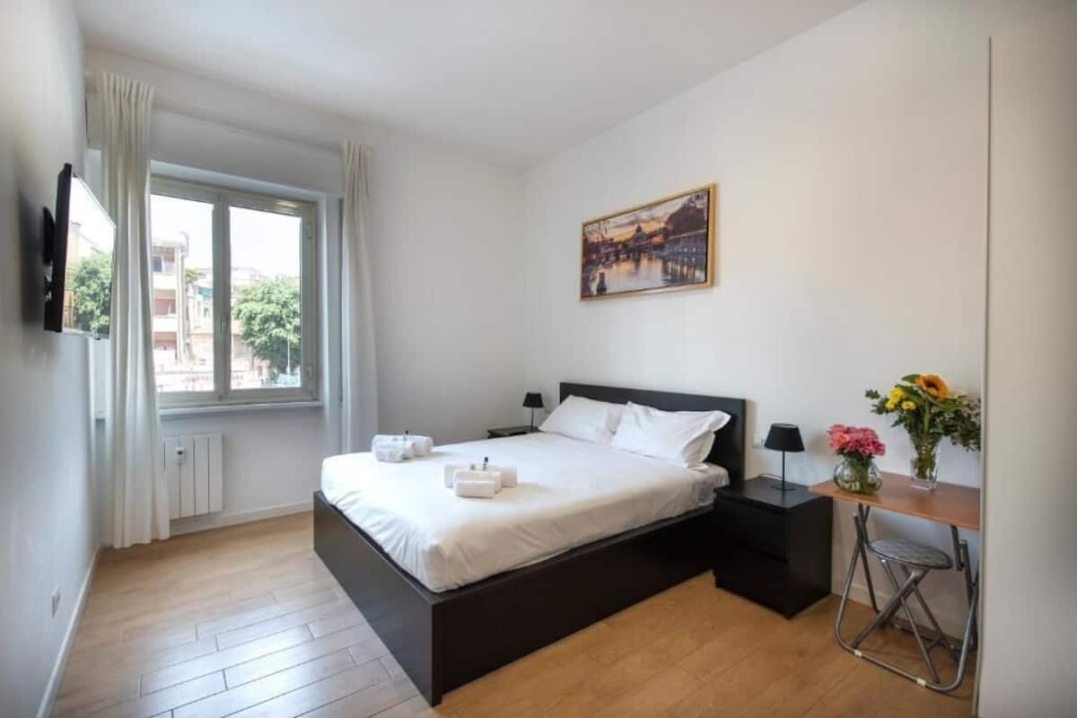 Un dormitorio con cama, mesitas de noche, un escritorio, una silla, una ventana con cortinas, un cuadro en la pared y un radiador.