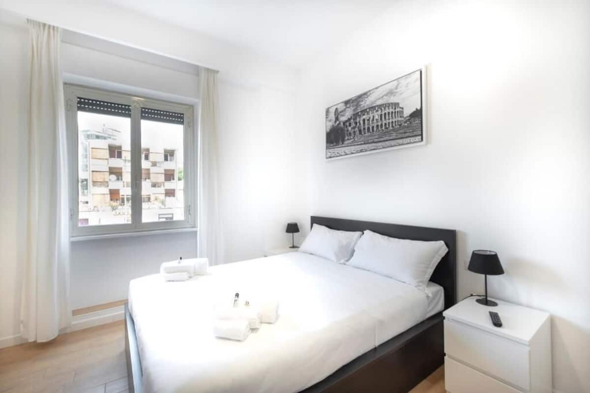 Habitación de hotel con cama, mesita de noche, lámpara, ventana con cortinas e imagen colgada en la pared.