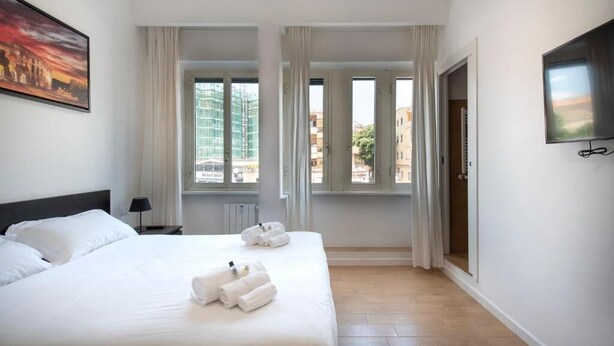 Habitación de hotel con cama, dos ventanas, un televisor y un cuadro en la pared.
