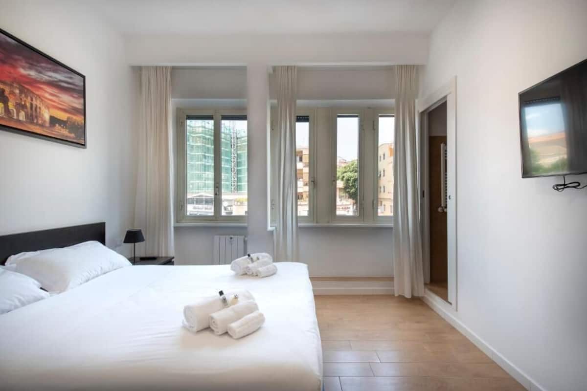 Habitación de hotel con cama, dos ventanas, un televisor y un cuadro en la pared.